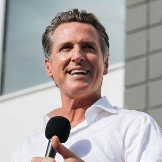 California Governor Gavin Newsom, wearing a white collared shirt, speaking into a microphone and looking to the left.